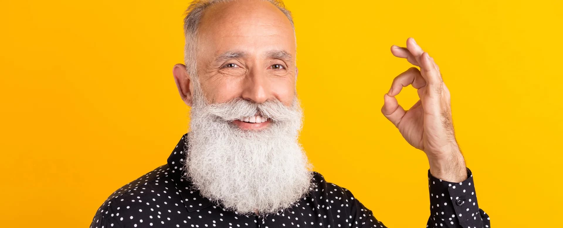 Älterer Mann mit langem weißen Bart und Punktenhemd zeigt lächelnd ein ‘Okay’-Handzeichen vor gelbem Hintergrund.