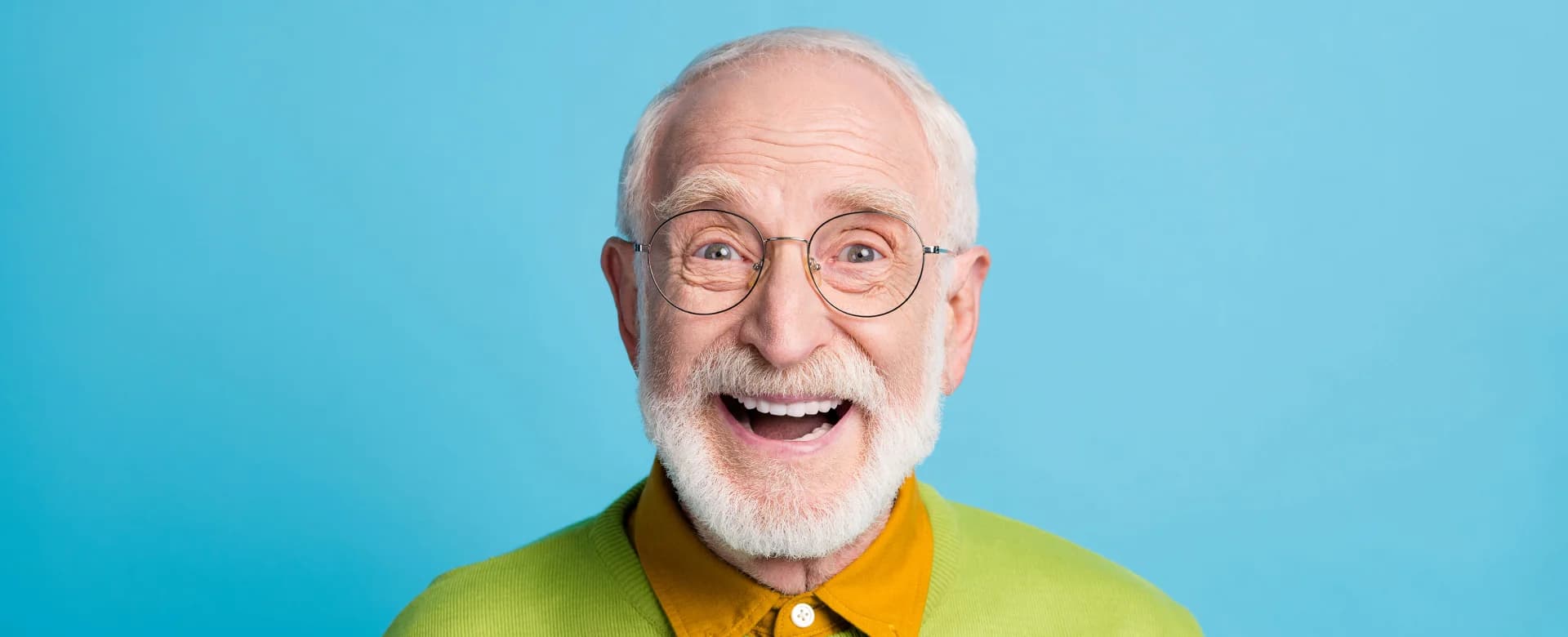 Fröhlich lachender älterer Mann mit weißem Bart, runder Brille und buntem Outfit vor hellblauem Hintergrund.