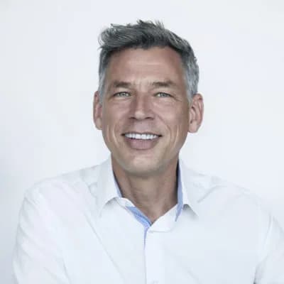 Ein Portraitbild. Er trägt ein weißes Hemd, hat grau meliertes Haar und lächelt in die Kamera. Der Hintergrund ist hell und neutral.