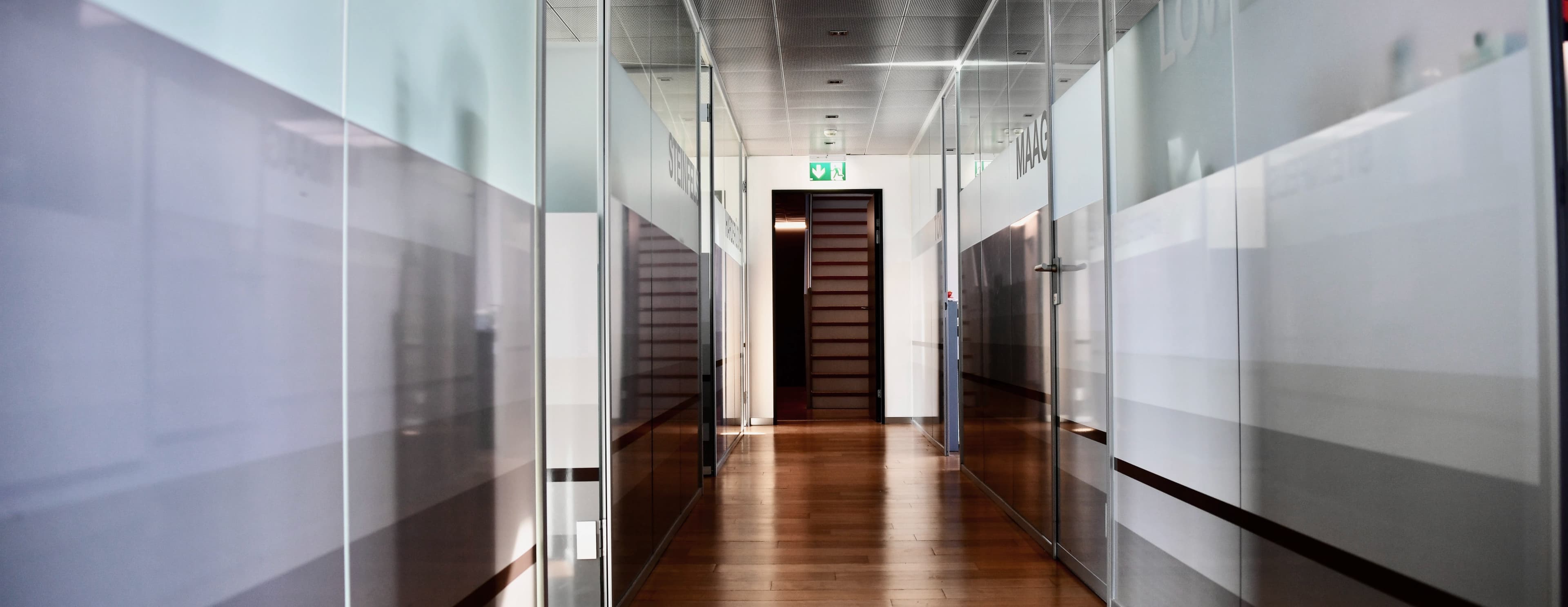 Flur der Swiss Dental Center Praxis mit Glaswänden und Raumbezeichnungen, Holzfußboden sowie gut sichtbarer Notausgangsbeschilderung an der Decke.