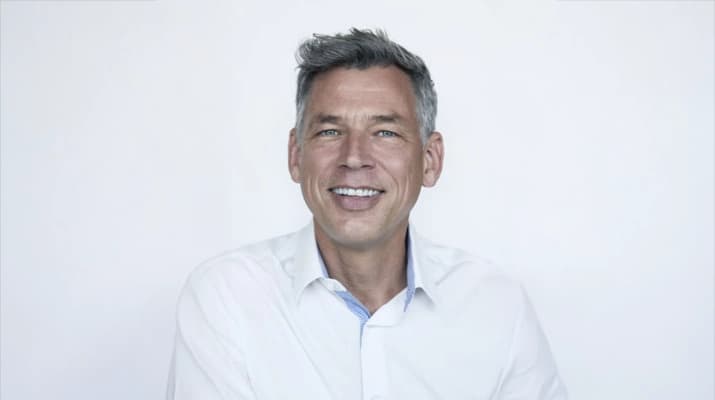Ein Portraitbild. Er trägt ein weißes Hemd, hat grau meliertes Haar und lächelt in die Kamera. Der Hintergrund ist hell und neutral.