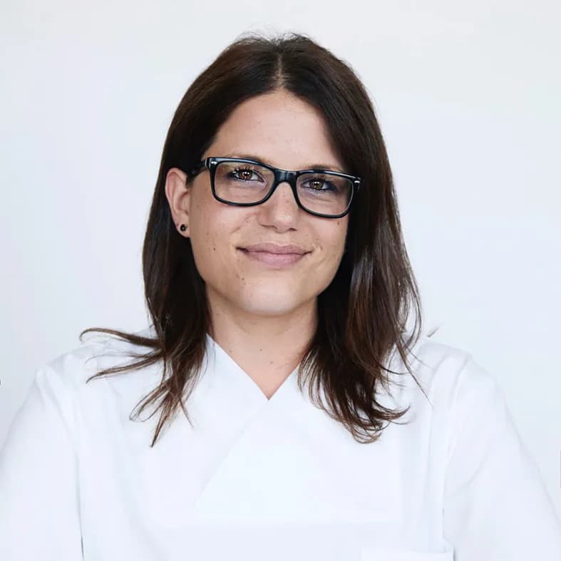 Sie trägt eine Brille und ein weisses Oberteil, ihr Blick ist offen und freundlich.