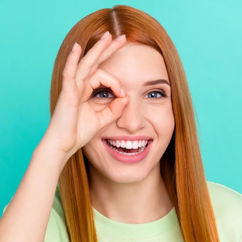 Junge Frau mit roten Haaren lächelt und formt mit Daumen und Zeigefinger ein ‘Okay’-Zeichen vor einem Auge, vor türkisfarbenem Hintergrund.
