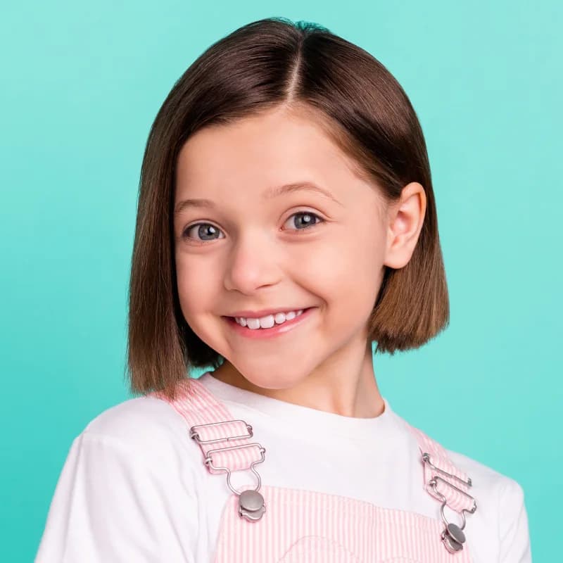 Lächelndes junges Mädchen mit braunem Bob-Haarschnitt und weißem Shirt mit rosa Trägern vor türkisfarbenem Hintergrund.