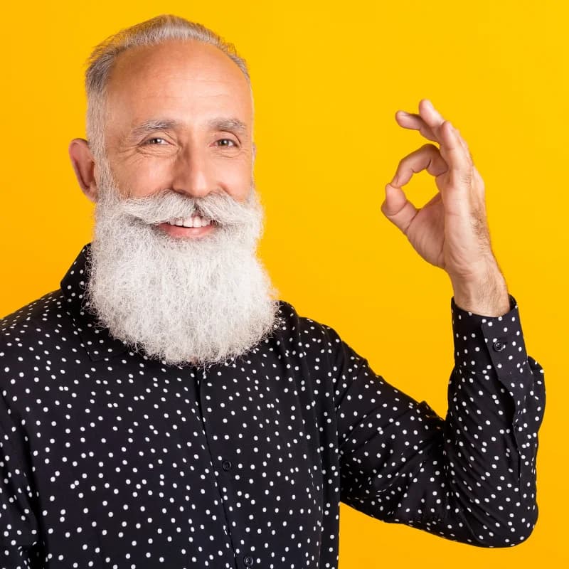 Älterer Mann mit langem weißen Bart und Punktenhemd zeigt lächelnd ein ‘Okay’-Handzeichen vor gelbem Hintergrund.