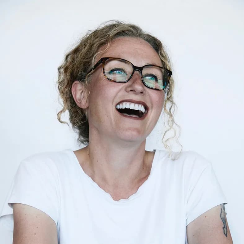 Sie lacht ausgelassen und trägt eine Brille sowie ein weisses Oberteil.